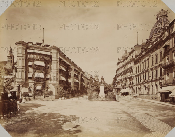 Leopoldplatz in Baden-Baden, album page, photograph, This photo is part of an album., inscription: '1673 Baden-Baden Leopoldplatz LL', photographer: LL, Baden-Baden, 1890 - 1899, Baden-Württemberg, paper, albumen print, albumendruk, height 258 mm x width 355 mm, square, circus, etc. (city(-scape) with figures, staffage), A wide city street flanked by multi-story ornate buildings with decorative facades and awnings stretches toward a central statue on a pedestal surrounded by low hedges and trees, the left building covered in climbing plants and window awnings and the right building featuring balconies and a large domed roof with patterned tiles, lamp posts and flagpoles rising above the rooftops, a cobblestone roadway and sidewalk in the foreground, a horse-drawn carriage and a few pedestrians visible near the left edge, and distant towers and architectural details visible down the street beneath a pale sky.
