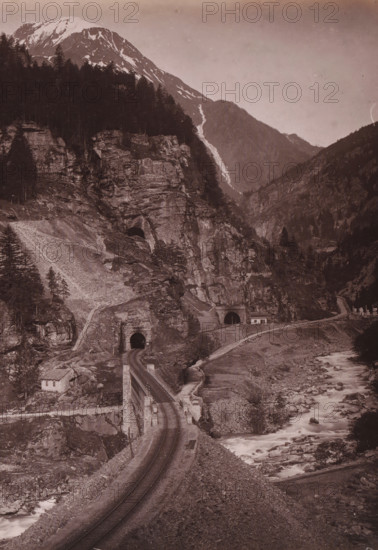 Gotthard Railway Faido, View of the Gotthard Railway at Faïdo, album page, photograph, This photo is part of an album., annotation: 'Gotthard railway at Faïdo', number: '12894', photographer: Giorgio Sommer, c. 1885 - c. 1892, paper, albumen print, height 199 mm x width 254 mm, railway, train, mountains, Faido, A sepia-toned landscape showing a rocky mountain slope with patches of snow near the peak, dense dark tree cover on parts of the slopes, and exposed rock faces. A river with white foamy water runs through a rocky riverbed alongside a narrow road or path that winds up the slope. Stone tunnels with dark arch openings are built into the rock next to a curved section of road or rail, and small stone structures or buildings sit near the road and tunnel entrances. The sky appears clear and pale above the mountains, and the image is framed by a light border along the top and side.