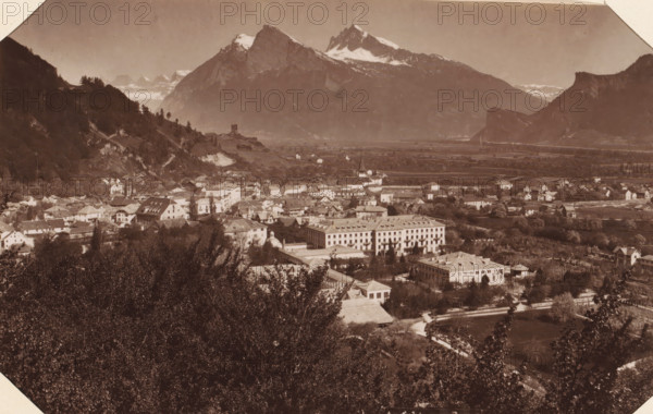 View of Ragaz, View of Bad Ragaz with mountains in the background, album page, photograph, This photo is part of an album., annotation: 'Ragatz - Panorama', c. 1885 - in or before 1892-05-02, photographer, paper, albumen print, height 134 mm x width 206 mm, city-view in general, 'veduta', mountains, Bad Ragaz, A wide sepia photograph shows a town nestled in a valley with clustered buildings of various sizes and roof shapes, including a few larger rectangular structures with regular window patterns and a small domed roof among them, surrounded by trees and gardens. In the foreground there is dense vegetation and leafy trees partially obscuring the lower part of the town. The middle ground features a mix of houses, roads, and open fields with a church steeple rising above nearby rooftops. Hills flank the town on both sides, with one side showing terraced slopes and scattered trees. In the background towering snow-capped mountains dominate the skyline under a clear sky.