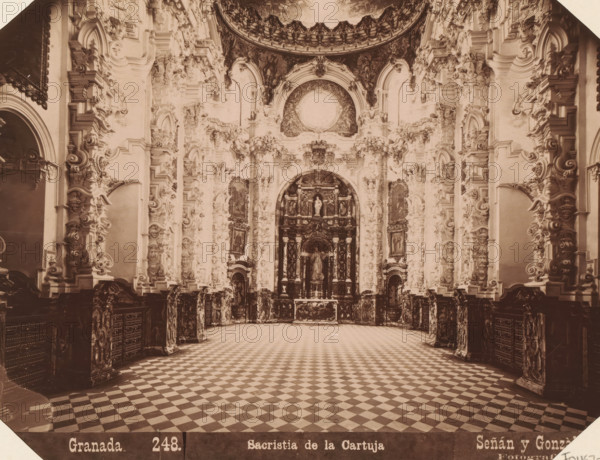 Interior of the sacristy of the Real Monasterio de La Cartuja in Granada, Granada. Sacristia de la Cartuja, album page, photograph, This photo is part of an album., annotation: 'Granada - Sacristie der Cartuga', number: '248.', photographer: Señan y Gonzalez, c. 1885 - in or before 1892-03-12, paper, albumen print, height 159 mm x width 208 mm, parts of church interior: sacristy, Andalusia, Granada, A large ornate interior space with a wide checkered floor leading toward a richly decorated altar niche, the walls and columns filled with elaborate carved and sculpted ornamentation including scrolls, floral motifs, and framed panels, high arched openings and recessed alcoves, a central arched area containing statues and layered architectural elements, decorative molding and relief work rising to a domed or curved ceiling with painted or darkened panels, dark base panels and carved cabinetry lining the lower walls, and soft sepia-toned lighting across the whole scene.