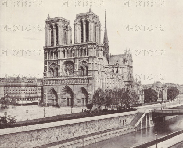 Notre-Dame of Paris, album page, photomechanical print, This print is part of an album., maker: Angerer & Göschl, C., 1880 - 1900, paper, autotype, height 201 mm x width 248 mm, river, church (exterior), Paris, Notre-Dame (Paris), A large Gothic cathedral façade dominates the image with two tall square towers, a central circular rose window, ornate arched doorways, and rows of vertical columns and sculptural details. A slender pointed spire rises behind the towers above the roofline. Trees and a low balustrade sit in front of part of the building, and a riverside embankment with stone steps, a retaining wall, and a bridge span the foreground. To the left and behind the cathedral are multi-story buildings with regular windows and mansard roofs, and lamp posts line the open paved area between the cathedral and the embankment. The scene is shown in a monochrome, grainy tone.