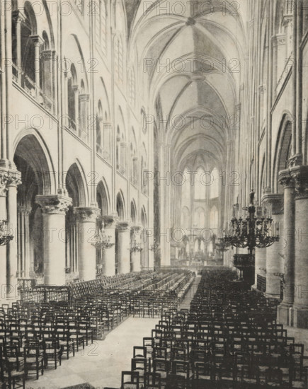 Interior of Notre-Dame in Paris, album page, photomechanical print, This print is part of an album., maker: Angerer & Göschl, C., 1880 - 1900, paper, autotype, height 250 mm x width 198 mm, interior of church, Paris, Notre-Dame (Paris), A large interior space with tall vaulted ceilings and a long central aisle lined with rows of wooden chairs, flanked on one side by a series of massive stone columns supporting rounded arches and an upper gallery with smaller arches and windows, and on the other side by similar columns and architectural details; ornate chandeliers hang from the ceiling and are positioned along the side aisles, decorative capitals sit atop the columns, and light filters through tall arched windows at the far end, illuminating the distant choir area and altar rail.