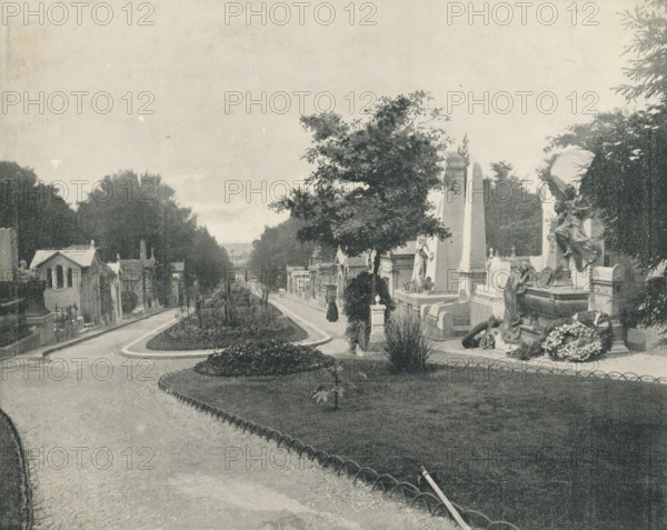 Père-Lachaise in Paris, album page, photomechanical print, This print is part of an album., maker: Angerer & Göschl, C., 1880 - 1900, paper, autotypie, height 201 mm x width 248 mm, necropolis, Paris, Cimetière du Père-Lachaise, A wide view of a tree-lined avenue with a curved pathway and a grassy central island, flanked by stone mausoleums and sculpted grave monuments, some topped with statues and ornate carvings; a few tall trees and shrubs rise among the memorials, and a person in dark clothing stands on the path near a large upright stone monument, while another sculpted figure reclines on a tomb surrounded by floral arrangements.