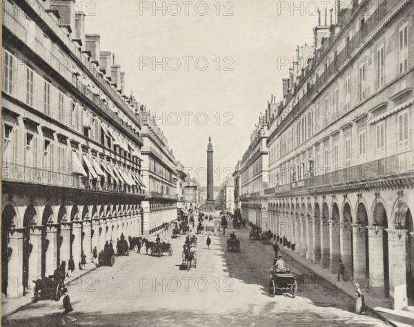 View of the Austerlitz Column, album page, photomechanical print, This print is part of an album., maker: Angerer & Göschl, C., 1880 - 1900, paper, autotype, height 249 mm x width 200 mm, historical persons, monumental adornments (in city), animal traction, animal-drawn vehicle (carriage etc.), Paris, A wide street flanked by long rows of multi-story buildings with repeating arches at ground level and shuttered windows above, a tall column topped with a statue visible in the distance at the center, horse-drawn carriages and riders moving along the street, pedestrians walking or standing near the sidewalks, decorative ironwork and awnings on some windows, and street lamps attached to the building façades.