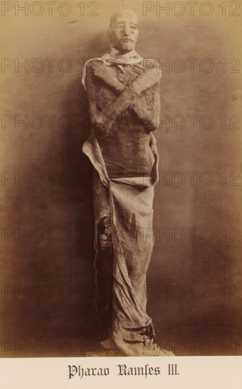 Mummy of Pharaoh Ramses III, Pharaoh Ramses III, photograph, page, inscription: 'Verlag von S. Mayer in Esslingen', publisher: Samuel M. Mayer, 1886, photographer, paper, albumen print, albumendruk, height 234 mm x width 164 mm, Person, Ramesses III (Pharaoh of Egypt), king, mummy, North Africa, Egypt, A tall upright wrapped figure stands against a plain background, the body covered in aged linen bandages and cloth that hang and fold in places, with arms crossed over the chest and portions of the torso and limbs exposed where the wrappings are worn; the face is partially covered with tight bandaging but shows facial contours and a closed mouth, and the lower cloth drapes down to the base with visible texture and frayed edges.
