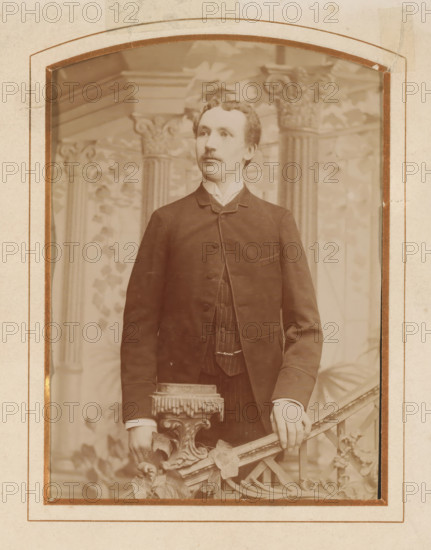 Portrait of a young man, standing by a stair railing, album page, photograph, cabinet photograph, This photo is part of an album., inscription: 'Frans E. Arvidsson, Helsingborg, Drottninggatan 18., Plåten förvaras, No... för efterbeställning.', photographer: Frans Edvard Arvidsson, 1872 - 1900, paper, pasteboard, albumen print, albumendruk, height 139 mm x width 98 mm, historical persons, standing figure, youth, adolescent, staircase, A sepia-toned portrait of a young man standing, shown from head to mid-thigh, wearing a dark suit jacket over a vest and a high-collared shirt; he has short wavy hair and a small mustache and looks slightly to his left, with one hand resting on an ornate carved railing that features decorative leaves and a fringed upholstered finial, and a painted backdrop behind him shows architectural columns and vine-like foliage; the photograph is mounted within a worn paper frame with rounded top corners and visible tape and discoloration along the edges.