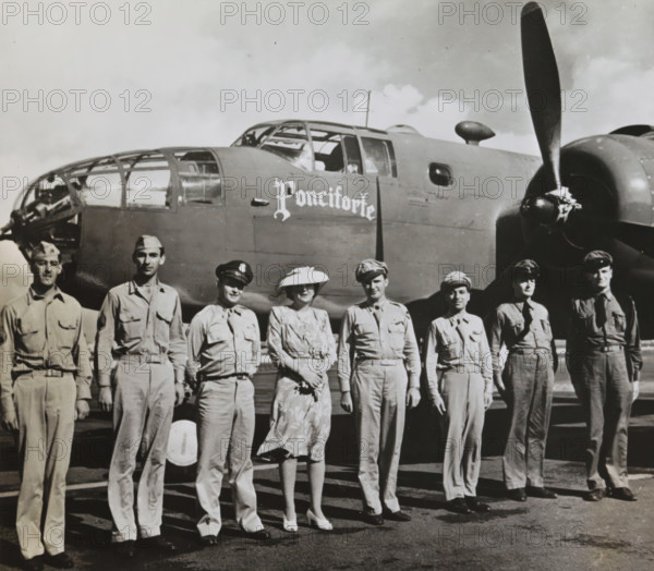 Princess Juliana with American aviators in front of an airplane, Princess Juliana with the crew of the American escorting plane, photograph, inscription: 'Princess Juliana's visit to Suriname...', stamp: 'copyright Netherland Government Information Bureau', Suriname, 1943, photographer, paper, gelatin silver print, gelatin silver print on photographic paper, height 180 mm x width 240 mm, Person, Juliana of the Netherlands, A row of uniformed men and one woman stand in front of a large military aircraft, the plane showing a glazed nose section, cockpit windows, a propeller and engine cowling, and a rounded fuselage; the woman wears a patterned dress and a wide-brimmed hat while the men wear military shirts, trousers and various caps, all visible from head to toe with shadows cast on the ground beneath them.