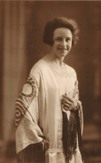 To Mr. E van Essen, Nora Janssens, Sept 1925 A'dam, Portrait of Nora Janssens, photograph, The photo is mounted on a passe-partout from photographer Geveke and placed in a folder., annotation: 'To Mr. E van Essen, Sept 1925 A'dam', signature: 'F. Geveke A'dam 10 Sarphatipark', photographer: Frits Geveke, Amsterdam, 1925, Frits Geveke, photographer, paper, gelatin silver print, developing gelatin silver print, height 173 mm x width 115 mm, height 332 mm x width 236 mm, Person, historical persons - BB - woman, Netherlands, A vintage sepia portrait of a smiling young woman with short wavy hair, wearing a light satin robe with patterned sleeves and long fringe, a lace undergarment visible at the neckline, and a ring on one finger as she lightly clasps the fabric at her chest, set against a plain softly blurred backdrop.