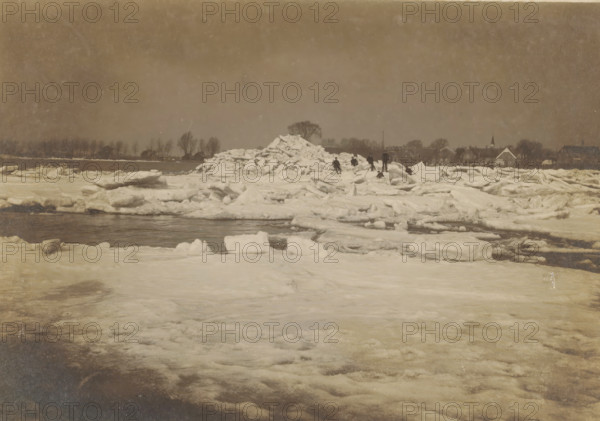 Ice floes in a river, Netherlands, photograph, check stamp: 'Atelier W. Visser Werkendam', photographer: W. Visser, Werkendam, 1910 - 1940, pasteboard, paper, gelatin silver print, developing gelatin silver print, height 120 mm x width 166 mm, height 239 mm x width 296 mm, river, drift ice, A wide frozen landscape with large sheets and chunks of ice piled and scattered across a snowy surface, a dark strip of open water cutting through the ice, several small figures standing and walking near a tall mound of broken ice, a distant shoreline lined with bare trees and a row of houses with pitched roofs and a steeple, a muted cloudy sky overhead.
