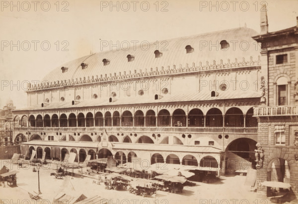 Market stalls in Padua, Padova. Il Salone, photograph, Padua, 1851 - 1900, photographer, pasteboard, paper, albumen print, albumendruk, height 125 mm x width 185 mm, height 144 mm x width 190 mm, market, square, circus, etc. - II - ideal city, A wide sepia photograph of a large multi-level stone building with rows of arches and an arched roof punctuated by small dormer openings; the facade shows decorative crenellations and circular windows above a long colonnaded gallery, with open archways on the lower level leading to covered stalls and awnings; in front of the building an open square contains market stalls with cloth canopies, carts, and scattered goods, and a solitary lamp post stands near the foreground; at the right edge a stone corner of another building with a balcony and sculpted figures is visible.