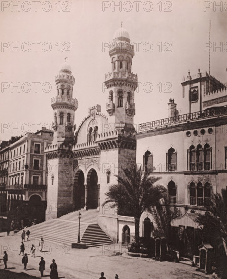 View of the Djamaa Ketchaoua and the Dar Hassan Pacha in Algiers, Alger, Cathédrale et Palais d'hiver du Gouverneur, photomechanical print, address: '...ue Bab-Azoun ALGER', number: '569', photographer: Alexandre Leroux, Algiers, cliché maker: anonymous, c. 1875 - c. 1900, Alexandre Leroux, photographer, cliché maker, North Africa, pasteboard, paper, collotype, photoprint, height 318 mm x width 246 mm, temple, shrine, Islam, Mohammedanism, palace, Djama'a Ketchaoua, Kasbah of Algiers, Dar Hassan Pacha, A large stone building with ornate arched doorways and wide steps leading up to an entrance dominates the scene, flanked by two tall cylindrical towers topped with domes and decorative balconies, and a façade featuring carved patterns and pointed crenellations; to the right, a connected structure displays arched windows with shutters, circular openings near the roofline, and an upper terrace with a balustrade, while palm trees rise in front of this section; on the left, a multi-story bui...