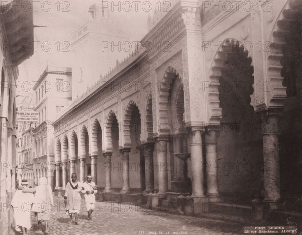 Rue de la Marine, View of the Djama'a al-Kebir in Algiers, photomechanical print, number: '117', address: '26 rue Bab-Azoun ALGER', photographer: Alexandre Leroux, Algiers, cliché maker: anonymous, c. 1875 - c. 1900, Alexandre Leroux, photographer, cliché maker, North Africa, pasteboard, paper, collotype, photoprint, height 247 mm x width 316 mm, temple, shrine, Islam, Mohammedanism, Djama'a al-Kebir, A narrow street scene with a row of tall, ornate arches supported by columns along one side of the street, each arch featuring scalloped inner edges and decorative panels above, and a series of buildings with multiple windows lining the opposite side; the street surface is cobbled and a small group of people wearing light-colored, loose garments and head coverings are walking and standing near the foreground, while the architectural details include carved motifs along the architrave, rounded column capitals, and a taller building or tower rising in the background.