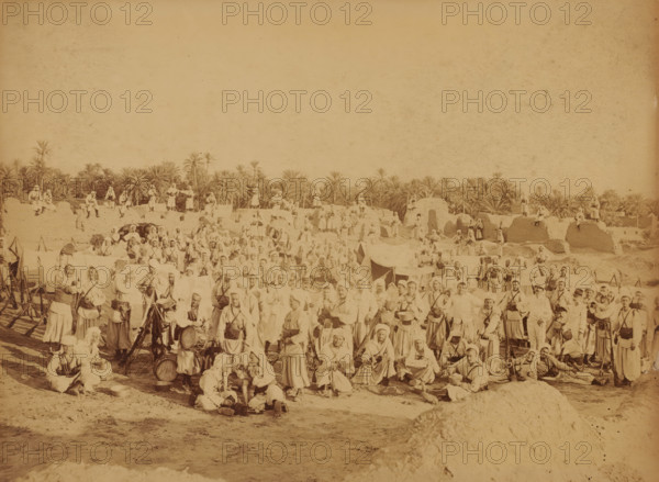 Fort Turc au Vieux Biskra, French garrison at Biskra, Algeria, Souvenir ... Biskra, photograph, inscription: 'Jul D...', Biskra, 1860 - 1900, photographer, North Africa, pasteboard, paper, albumen print, albumendruk, height 182 mm x width 241 mm, garrison, A wide sepia photograph showing a large group of people gathered outdoors with palm trees and low mud structures in the background; many individuals wear light-colored robes and head coverings, some hold musical instruments or drums, and others carry rifles or stand beside tripods and equipment; tents and makeshift shelters are scattered among the crowd, with people seated, standing, and leaning on the ground across the foreground and middle distance; the scene extends horizontally with figures positioned on earthen mounds and walls, and taller palm trees rising behind the settlement under an empty sky.