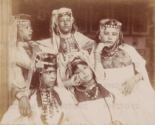 Group portrait of five unknown Ouled Naïl women in Algeria, Alger, groupe de filles d'Ouled Nail à Biskra, photograph, Algeria, 1890 - 1930, photographer, North Africa, paper, pasteboard, baryta paper, daylight gelatin silver print, height 247 mm x width 317 mm, anonymous historical persons depicted in a group, in a group-portrait - BB - woman, Five seated individuals wearing layered garments and headpieces decorated with round metal discs and dangling chains are shown in a close group portrait; some head coverings include veils and braided or beaded adornments, and several necklaces and bracelets with similar round metal decorations are visible on their chests and wrists; one person rests a hand under their chin while another leans their head on a hand, and their faces display neutral expressions against a plain, darker background.