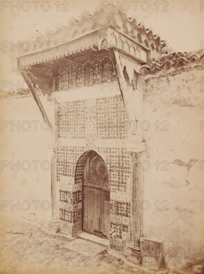 Tlemcen, Portal in Tlemcen, Algeria, photograph, number: '235', inscription: 'Morocco', 1860 - 1900, photographer, North Africa, pasteboard, paper, albumen print, albumendruk, height 279 mm x width 215 mm, closed door, A weathered doorway set into a tiled wall with ornate geometric patterns is visible, featuring an arched wooden door with decorative carved panels and metal ring handles; flanking the doorway are textured columns and a carved lintel, and above it a projecting wooden balcony with carved decorative brackets and an overhanging tiled roof supported by angled wooden beams; the surrounding plastered wall shows areas of wear and discoloration, and the foreground shows a cobbled street surface leading up to the entrance.