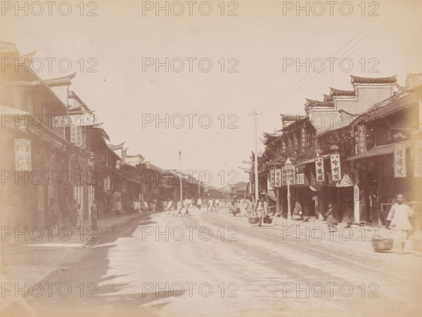 Street view of Nanking Road in Shanghai, photograph, annotation: 'Shanghai-Nanking road', Shanghai, 1850 - 1900, photographer, paper, pasteboard, albumen print, height 225 mm x width 310 mm, street (city(-scape) with figures, staffage), A wide street scene with rows of multi-story buildings on both sides featuring ornate upturned roof edges and hanging rectangular signs, a mostly empty upper sky above, lamp posts and a few utility poles with overhead wires, numerous blurred figures walking and standing along the street and sidewalks, some carrying baskets or goods, awnings and shopfronts lining the sidewalks, and a textured road surface stretching into the distance.