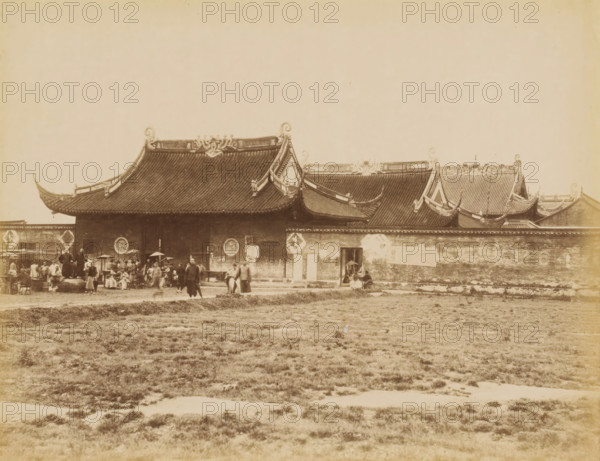 View of a city wall and residences in Shanghai, photograph, annotation: 'Shanghai-Chinese residences', Shanghai, 1850 - 1900, photographer, paper, pasteboard, albumen print, height 235 mm x width 310 mm, city-walls (city(-scape) with figures, staffage), A wide sepia photograph shows a long traditional building with sweeping tiled roofs and ornate upturned eaves, decorative ridge ornaments, and circular reliefs on the upper wall; a low wall runs along the front with a small doorway where a person stands partially sheltered by an umbrella, and a group of people gathered near the left side include figures standing, sitting, and carrying baskets and umbrellas; in front of the building a grassy open area stretches across the foreground.