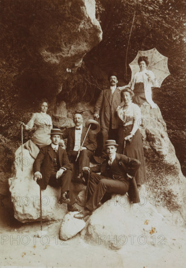 Group portrait of a tourist group in Valkenburg on a rock slope, Memory of Valkenburg L., photograph, Photo on passe partout with the name of the photographer printed on it., annotation: 'Memory of Valkenburg L.', signature: 'Jos. Crolla-Falise photographer Valkenburg (L.)', photographer: Jos. Crolla-Falise, Valkenburg (Limburg), 1890 - 1920, paper, daylight collodion silver print, height 168 mm x width 118 mm, height 255 mm x width 203 mm, anonymous historical persons depicted in a group, in a group-portrait, A sepia-toned photograph showing a group of well-dressed people posed on and around large rough rocks at the mouth of a shaded rocky overhang, with foliage and tree branches visible above; women in high-collared blouses and long skirts stand and sit among men wearing suits, ties, and hats, one woman holding a patterned parasol, several individuals holding walking sticks or canes, and facial hair visible on some of the men; the rock surfaces are textured and uneven, and the ground...
