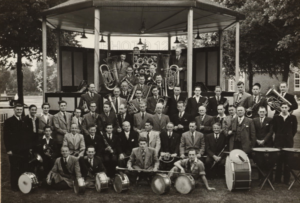 Group portrait of the 'Crescendo' harmony orchestra, photograph, photographer: NV Vereenigde Fotobureaux, Amsterdam, 1949, paper, gelatin silver print, gelatin silver print on photographic paper, height 120 mm x width 174 mm, anonymous historical persons portrayed in a group, in a group-portrait, banner, standard (as symbol of the state, etc.), musical instrument, group portrait, A large group of men posed together in front of a wooden bandstand, many wearing suits and ties and some in jackets, arranged in rows with brass and woodwind instruments visible throughout; several tubas and horns are positioned behind and beside the group, while trumpets, trombones, clarinets, and saxophones are held by seated and standing members; a row of drummers with snare drums and a bass drum sits at the front, with drumsticks and a drum major’s mace visible; the bandstand has a pitched roof supported by posts and hanging lamps, and decorative panels at the rear of the platform; trees and parkland are v...