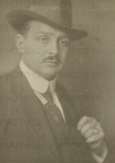 Portrait of the photographer Jacob Merkelbach, photograph, name: 'Bern. F.Eilers Amsterdam', photographer: Bernard Eilers, Amsterdam, 1910 - 1930, paper, daylight collodion silver print, height 230 mm x width 171 mm, height 407 mm x width 306 mm, Person, Jacob Merkelbach, portrait, self-portrait of photographer, A sepia-toned portrait of a man wearing a dark fedora and a suit jacket with a light-colored shirt and a tie, his face turned slightly toward the camera showing a thin mustache and a contemplative expression, one hand visible in the lower right holding an object, with a softly textured background and visible surface wear on the photograph.
