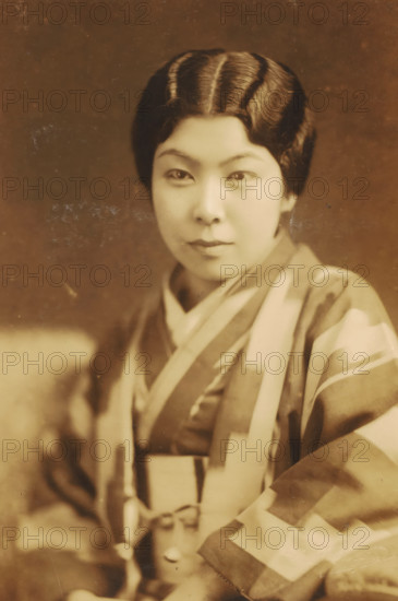 Portrait of an unknown Japanese woman, photograph, address: 'YOKOYAMACHO.HACHOJI., TEL. 320.', photographer: T Jingu, Tokyo, c. 1900 - c. 1920, Kanto, paper, pasteboard, baryta paper, daylight gelatin silver print, height 133 mm x width 89 mm, height 242 mm x width 156 mm, adult woman, anonymous historical person depicted, Japan, A sepia-toned portrait of a person wearing a patterned garment with broad stripes and geometric shapes, showing a close-up of the upper body and face. The person has neatly styled, dark hair with visible waves and a center part, and a smooth, even complexion. The eyes are looking forward, the eyebrows are softly curved, and the lips are closed. The garment has layered collars and draped fabric across the shoulders and chest, with a tied sash visible at the waist. The background is plain and softly textured, and the image has small surface marks and scratches.