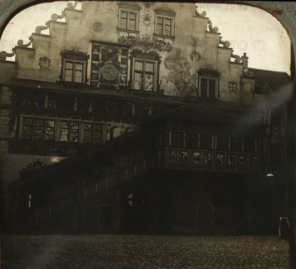 Rear facade of the Lindau town hall, Rathhaus in Lindau, Hinterseite, stereograph, number: '759.', Lindau im Bodensee, 1900 - 1940, photographer, Bavaria, paper, plastic, pasteboard, organic material, cellulose nitrate film, gelatin silver print, developing gelatin silver print, height 75 mm x width 150 mm, town hall, rear facade, A monochrome view of an ornate building façade with decorative gables and patterned windows, featuring a large circular clock framed by sculpted reliefs and coats of arms, painted murals or frescoes on the plastered wall, a projecting wooden gallery with carved panels and small leaded glass panes, an external staircase with a balustrade leading to the upper level, a cobbled foreground, and an arched decorative border at the top and sides of the image.