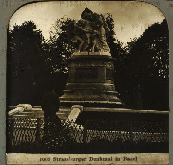 Strassburger monument in Basel, Strassburger Denkmal in Basel, stereograph, number: '1002', Basel, after sculpture by Frédéric Auguste Bartholdi, 1900 - 1940, Frédéric Auguste Bartholdi, photographer, after sculpture by, paper, plastic, pasteboard, organic material, cellulose nitrate film, gelatin silver print, developing gelatin silver print, height 75 mm x width 150 mm, monument, statue, A stone monument with a sculpted group on top stands amid trees and dark foliage, the sculpture depicting draped figures in flowing garments and wings on a raised pedestal with carved panels and decorative molding; stone steps and a low railing lead up to the base where a person in dark clothing faces the monument and another person stands to the side; a decorative fence and plants occupy the foreground and the sky above is pale.