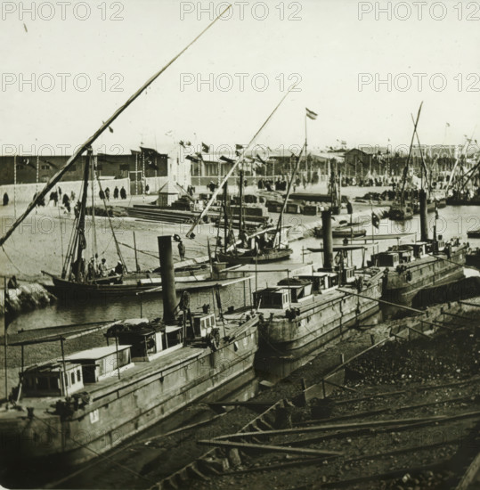 Inauguration of the Suez Canal. Ismaïlia, canal d'eau douce, arrival of the scheiks arabes (instantanee), Arrival of the Arab sheikhs for the opening of the Suez Canal, Egypt, stereoscopic photograph, number: '10011', photographer: Ferrier Père-Fils et Soulier & Léon et Lévy, Suez Canal, 1869, Ferrier Père-Fils et Soulier & Léon et Lévy, photographer, North Africa, inorganic material, glass, slide, height 84 mm x width 171 mm, canal, A waterfront scene showing multiple moored boats with tall masts and rigging alongside a quay, wooden decks and small cabin structures on the boats, and piles of cargo and crates on the shore; a row of larger vessels extends into the distance with visible chimneys and winches, and smaller rowing boats are tied near the waterline; long spars and ropes cross the foreground and middle ground, and people are visible on some boats and walking along the quay; beyond the quay a low wall separates the water from an open area with stacked materials, tents or tempor...