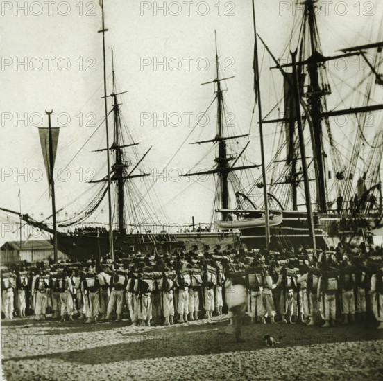 Soldiers in the port of Port Saïd for the opening of the Suez Canal, Egypt, Inauguration du Canal de Suez. Embarquement des troupes à Port-Saïd (instantanee), stereoscopic photograph, number: '10007.', photographer: Ferrier Père-Fils et Soulier & Léon et Lévy, Port Said, 1869, North Africa, inorganic material, glass, slide, height 84 mm x width 171 mm, harbour, troop movements, transportation, A crowded scene at a dock with tall ship masts and rigging rising into the sky behind a large group of uniformed people standing in formation, many seen from the back wearing light-colored trousers and carrying packs on their shoulders, while a single person stands facing them in the foreground and a small dog sits nearby on the ground; the ships show multiple spars, ropes, lifeboats, and hull details, and a tall flagpole with a crescent-topped finial appears at the left edge.