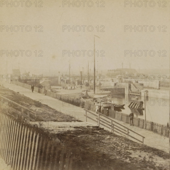 View of the harbor and locks of IJmuiden, IJmuiden, Collection A, Geographical sculpture gallery, collected by MJ Broekhuijsen, headmaster of a school in Amsterdam, stereograph, inscription: 'We are standing to the SE of the harbor. A boat is currently being locked through the lock.', number: 'A.5.', number: '5.', inscription: '58', publisher: Merkelbach & Co., Amsterdam, compiler (person in literature): Martien Johan Broekhuijsen, 1900 - 1902, photographer, compiler (person in literature), pasteboard, paper, albumen print, height 88 mm x width 177 mm, harbor, lift-lock, canal lock, A faded sepia scene showing a waterfront area with a boardwalk and piers, wooden fences running along a sloped embankment, small boats moored near the berths, tall masts and flagpoles rising from the docks, indistinct figures walking along the pathway and standing near the railings, a large pier structure extending into the water in the distance, and weathered ground and patterned shadows in the foreground.