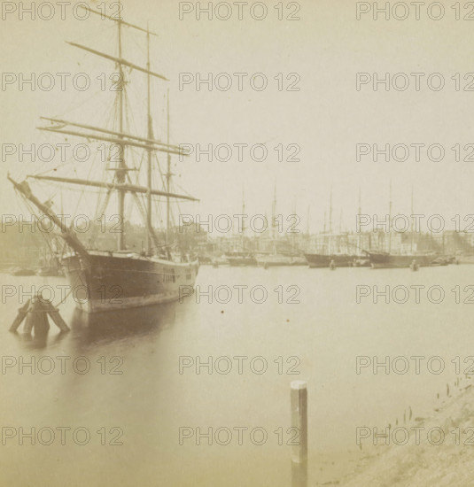 View of a city and a harbor with a ship in the foreground, Hollande, stereograph, Netherlands, 1873 - 1890, photographer, pasteboard, paper, albumen print, height 87 mm x width 177 mm, harbor, prospect of city, town panorama, silhouette of city, ships (in general), A faded sepia photograph showing a harbor scene with a large wooden sailing ship in the foreground at left, its tall masts, yards, and rigging visible, and the ship's bow facing slightly toward the viewer; a series of smaller masts and hulls of other vessels extend across the midground and recede into the distance, and a waterfront with low buildings is seen behind them; calm water fills the lower half of the image, reflecting the vessels, and a single wooden post rises from the water near the center-right; the overall image has a soft, aged texture and muted tones.