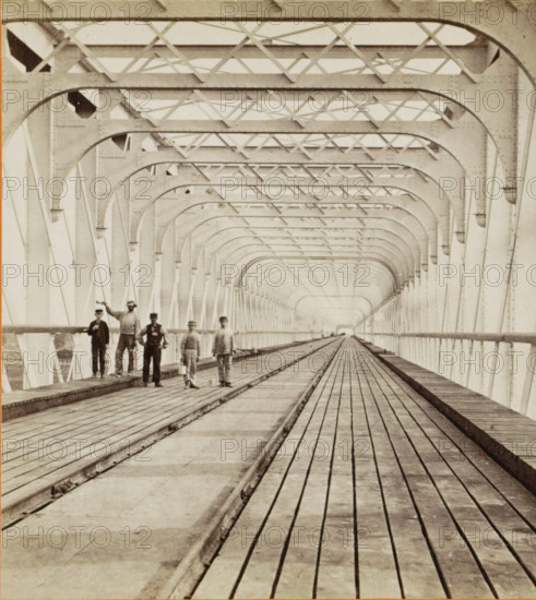 Kuilenburg railway bridge over the Lek, stereograph, photographer: Pieter Oosterhuis, Culemborg, before 1868-09-06, pasteboard, paper, albumen print, albumendruk, height 86 mm x width 176 mm, bridge in village across river, canal, etc. - II - ideal landscapes, Kuilenburg railway bridge, A long covered bridge interior with repeating metal arches and cross braces forming a tunnel-like perspective, wooden planks running lengthwise along the floor with a central track, railings along the sides, and a small group of men standing on the left side near the foreground wearing period clothing and hats.