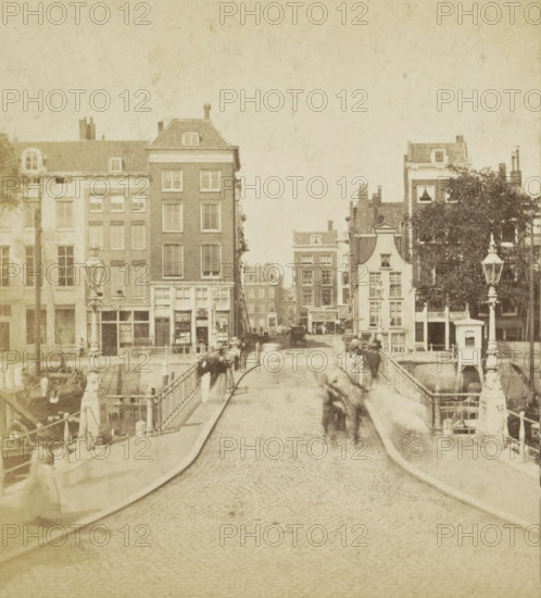 View of a bridge in Rotterdam, Leeuwen Bridge, Rotterdam, stereograph, inscription: '1200', 1861 - 1870, photographer, pasteboard, paper, albumen print, height 86 mm x width 175 mm, urban housing, bridge in city across river, canal, A cobblestone street runs from the foreground toward a bridge and a row of multi-story brick and plaster buildings, with tall narrow windows and sloped tiled roofs; ornate lamp posts and decorative railings line the bridge edges; indistinct blurred figures and horse-drawn carriages appear on the bridge and near the buildings; a large tree with dense foliage stands near one building on the right; canal water and moored boats are visible along the sides of the bridge; the overall image has a sepia tone and a slightly faded, aged appearance.