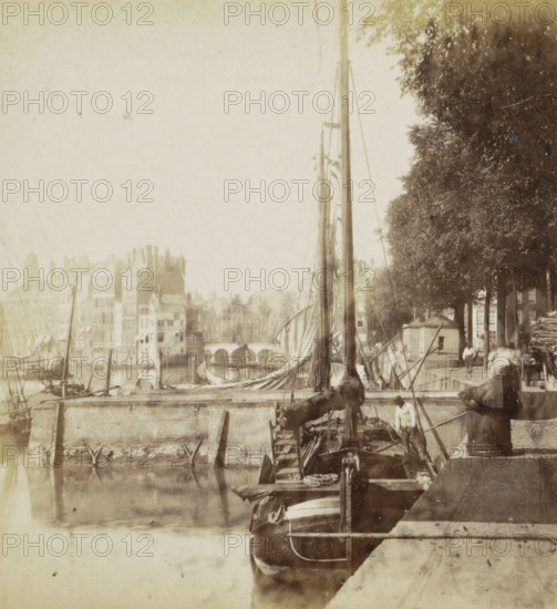 Cityscape with a harbor in Rotterdam, stereograph, Rotterdam, 1861 - 1870, photographer, pasteboard, paper, albumen print, height 75 mm x width 150 mm, harbor, city-view in general, 'veduta', ships (in general), A riverside scene with boats moored along a stone quay, tall masts and rigging rising above a vessel with rolled sails, a figure standing on one of the boats near the stern, another figure seated on the quay edge, clustered buildings with varied rooflines visible across the water, an arched bridge in the distance, and a row of leafy trees lining the quay casting dappled shade.