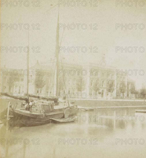 View of the Academic Hospital on the Catharijnesingel in Utrecht, The City Hospital., L'hopital., Das Krankenhaus., The Hospital, Utrecht, stereograph, inscription, 1871 - 1890, photographer, pasteboard, paper, albumen print, height 88 mm x width 178 mm, canals, waters (in city), hospital, ships (in general), Catharijnesingel, A pale, faded photograph showing a waterfront scene with several moored boats in the foreground, masts rising vertically and rigging visible, a small dinghy tied alongside, and the water reflecting the vessels; behind the boats a multi-story stone building with arched windows, decorative cornices, and a central pediment is visible across a paved area lined with small trees.