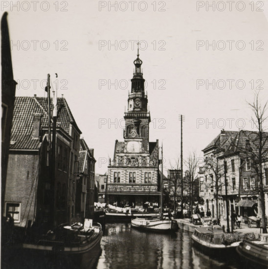 View of the Waag in Alkmaar, De Waag, Die Stadwage, The weight of the town Alkmaar, La balance de la ville Alkmaar, Alkmaar, stereograph, number: 'No. 2662.', number: 'R. 2662', 1900 - 1935, photographer, paper, gelatin silver print, developing gelatin silver print, height 89 mm x width 180 mm, city-view in general, 'veduta', weigh-house, Waag (Alkmaar), A narrow canal runs toward a multi-story building with an ornate clock tower rising above the rooftops, the tower topped by an openwork lantern and spire; moored boats fill the canal in the foreground with reflections on the water; rows of gabled buildings line both sides of the canal with tiled roofs and narrow facades; people are visible along the quays and near market stalls, and leafless trees stand between the buildings on the right side of the scene.