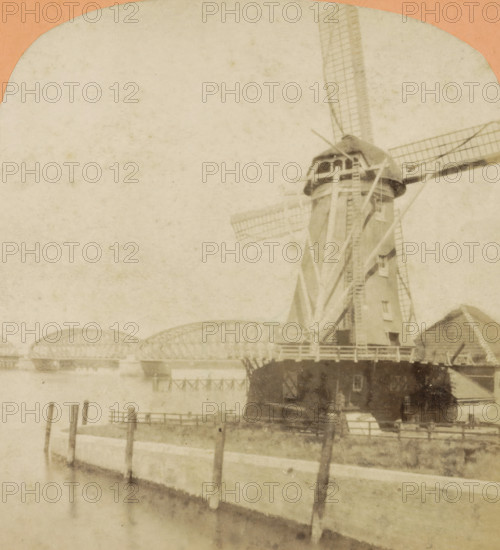 Vue prize a Dordrecht, View of a mill and the railway bridge over the Oude Maas near Dordrecht, Hollande pittoresque, stereograph, number: '2293.', photographer: Jules Hippolyte Quéval, Dordrecht, 1873 - 1890, pasteboard, paper, albumen print, height 86 mm x width 178 mm, windmill in landscape, bridge, Spoorbrug, A faded sepia scene showing part of a large windmill with its tower, cap, and lattice blades visible on the right side, a wooden railing and platform around its base, adjacent pitched roofs, a calm body of water in the foreground with wooden posts and a low concrete or stone quay, and a distant arched metal bridge spanning the water toward the left, all under a pale, textured sky.