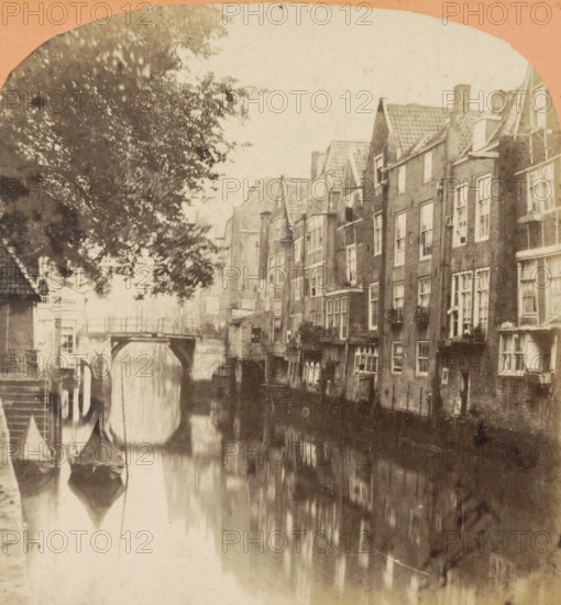 Dordrecht. Vue prize sur le vieux canal, View of a harbor in the city center of Dordrecht, Hollande pittoresque, stereograph, inscription: '40', number: '2284 bis.', photographer: Jules Hippolyte Quéval, Dordrecht, 1873 - 1890, pasteboard, paper, albumen print, albumendruk, height 87 mm x width 178 mm, city-view in general, 'veduta', canals, waters (in city), A narrow canal bordered by tall brick buildings with multiple windows and steep tiled roofs runs through the scene, their facades reflected in the water below; small boats with pointed covers float near a stepped stone bank on the left, and a low arched bridge spans the canal toward the center; a large tree with dense foliage overhangs the left side, casting mottled shadows, while architectural details like chimneys, window boxes, and small external stairways are visible along the row of houses.