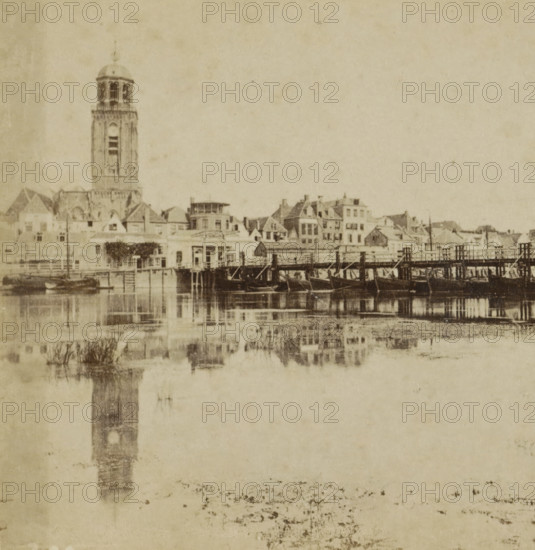 Deventer, View of the IJssel, the pontoon bridge and the Grote or Lebuïnus Church in Deventer, Collection A, Geographical sculpture gallery, collected by MJ Broekhuijsen, headmaster of a school in Amsterdam, stereograph, inscription: 'seen from the opposite side of the IJssel. Ship bridge, with a drawbridge on the city side.', number: '20.', publisher: Merkelbach & Co., Amsterdam, compiler (person in literature): Martien Johan Broekhuijsen, 1900 - 1902, photographer, compiler (person in literature), pasteboard, paper, albumen print, height 86 mm x width 176 mm, city-view in general, 'veduta', parts of church exterior and annexes: tower, pontoon bridge, Grote or Lebuinuskerk, A sepia-toned waterfront scene showing a row of buildings and houses along a shoreline with a tall tower rising among them, the tower featuring a domed top and an open belfry area, and smaller peaked and flat roofs surrounding it; a wooden pier or bridge extends across the water in front of the buildings with boats...