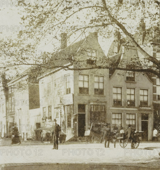 Street scene in The Hague, Hague, stereograph, photographer: Wilhelm Frederick Antonius Delboy, The Hague, 1884 - 1928, paper, pasteboard, daylight collodion silver print, height 88 mm x width 179 mm, urban housing, street, A street scene with tall narrow buildings showing multiple windows and pitched roofs partially obscured by tree branches in the foreground, a lamppost near a building entrance, a few horse-drawn carts parked by the curb, and several people standing or walking near the carts and building entrances.