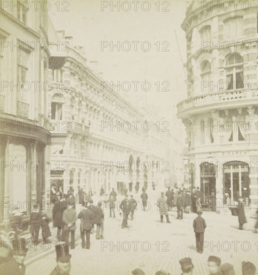 View of the Korte Poten in The Hague, The Hague, Korte Poten, stereograph, 1861 - 1870, photographer, pasteboard, paper, albumen print, albumendruk, height 88 mm x width 180 mm, urban housing, street, A faded street scene showing tall ornate buildings with arched windows and decorative facades lining both sides of the thoroughfare, a rounded corner building with a balustraded balcony visible on the right, shopfronts and window displays at street level, numerous people walking and standing along the street wearing long coats and hats of various styles, some clustered near shop entrances and others scattered down the roadway, a few figures nearer the foreground with top hats, a row of narrow dark trees or tall shrubs receding into the distance along the center of the street, and a pale sky above.