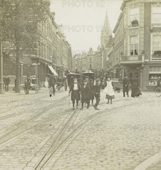 s Gravenhage.- Prinsenstraat, View of the Prinsestraat in The Hague, stereograph, number: '72.', The Hague, 1882 - 1920, photographer, paper, pasteboard, gelatin silver print, developing gelatin silver print, height 87 mm x width 177 mm, urban housing, street, Prinsestraat, A cobblestone street with visible tram tracks runs toward a distant church spire flanked by rows of multi-story brick buildings; people in period clothing walk and stand in the street, including a group of boys in the foreground and a woman in an apron; horse-drawn carriages and a bicycle appear among the pedestrians; shopfronts and awnings line the buildings, some with window curtains and small balconies; a large tree stands on the left sidewalk and a handcart is visible near the curb.