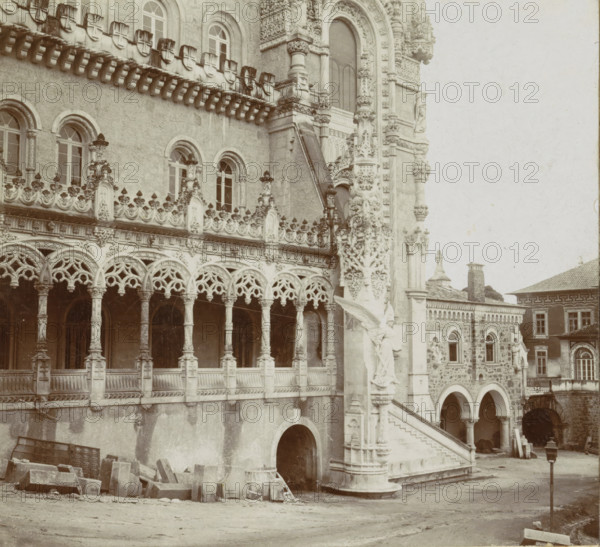 Domaine Royale du Bussaco. Façade principale de l'Hôtel, Facade of the Palácio Hotel do Buçaco, stereograph, inscription: 'Photographia Conimbricense', photographer: José Maria dos Santos, Palácio Hotel do Buçaco, 1869 - 1900, José Maria dos Santos, photographer, paper, pasteboard, baryta paper, gelatin silver print, developing gelatin silver print, height 88 mm x width 178 mm, palace, A detailed stone building facade with ornate carved arches and columns, a row of arched windows with decorative frames, and a balustraded gallery featuring intricate lace-like stonework; a sculpted stairway with carved figures and reliefs leads up beside a column richly decorated with floral and geometric motifs, while lower arches open into shadowed passageways and scattered stone blocks and masonry lie on the ground in front; adjoining structures with patterned stone walls, arched openings, and rectangular windows are visible at the side, and a lone lamp post stands in the courtyard area.