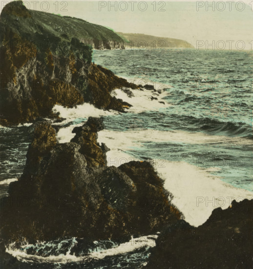 Bornholm Löwenklikken, Kamelhovederne og Løvehovederne, rocks in the shape of camel and lion heads, Bornholm, stereograph, monogram: 'BW', number: 'S 24984', inscription: 'Dänemark. Bornholm. Löwenklippen., Denmark. Bornholm. Lion-cliffs., Denmark. Bornholm. Ecueils de lion., Dinamarca. Bornholm. Escollos de león.', photographer: BW, 1898 - 1935, paper, baryta paper, gelatin silver print, hand coloring, developing gelatin silver print, height 89 mm x width 168 mm, rock-formations, A rocky coastline with dark jagged cliffs extending into a choppy sea, waves breaking against the rocks and creating white foamy surf, the water showing variations of blue and green, a distant headland visible under a pale sky, and textured rock faces with patches of lighter and darker tones.