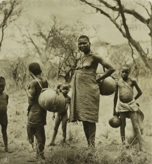 Kamba-Frau geht mit ihren Kindern Wasser holen, Vouw and children of the Kamba people fetch water, stereograph, number: 'S 6003', monogram: 'NPG', Africa, publisher: Neue Photographische Gesellschaft, Berlin, 1898 - 1935, photographer, paper, baryta paper, gelatin silver print, developing gelatin silver print, height 88 mm x width 180 mm, mother and child(ren), woman and child(ren) (family group), A sepia-toned photograph showing a group of people standing outdoors among leafless trees and dry grass, with a central adult woman wearing a wrapped cloth around her waist and chest, a necklace, and a strap over one shoulder, her hand resting on her hip and a large rounded container hanging from her other arm, several children around her also carrying rounded containers suspended by straps across their bodies, the figures shown from head to calf with bare feet and minimal clothing, the background filled with tangled branches and an open sky.