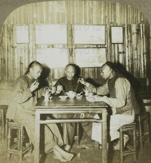 Chinamen eating chow in a restaurant. Canton, China, Three Chinese men at a meal, stereograph, number: '168', copyright notice: 'Copyright 1902 by CH Graves', number: '2', photographer: Carleton Harlow Graves, Guangzhou, publisher: Universal Photo Art Company, Philadelphia (possibly), 1902, North America, paper, pasteboard, baryta paper, gelatin silver print, developing gelatin silver print, height 89 mm x width 178 mm, eating, Three men sit around a wooden table in a dim interior, each holding chopsticks and reaching toward a central bowl of food; the man on the left is turned slightly toward the center, wearing a loose garment and holding food near his mouth, the man in the middle faces forward with a neutral expression and a simple dark shirt, and the man on the right leans in from the side wearing a vest over a long-sleeved shirt. The table holds bowls, small plates, a teapot, and two lanterns, and the men's feet are visible beneath the table, one pair bare and resting on the floor...