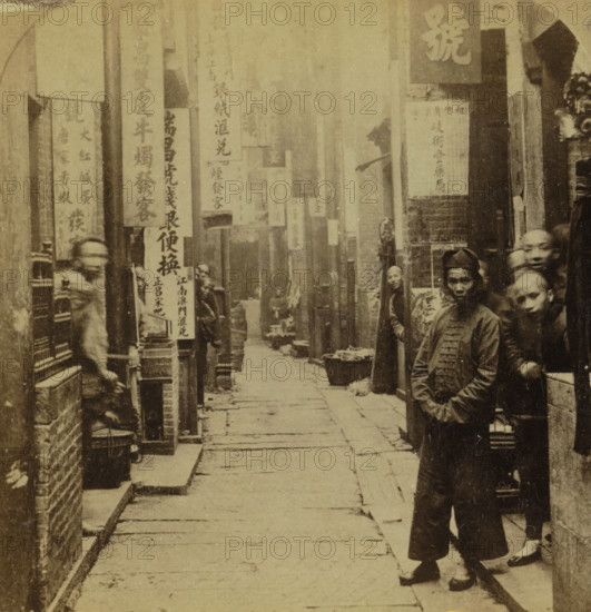 View of a Street in Canton, Geographical gallery of images, collected by MJ Broekhuijsen, headmaster of a school in Amsterdam, Collection H, stereograph, number: '6', number: '3', inscription: 'Canton: The first port opened in China for Europeans. Three times, as many inhabitants as Amsterdam. Narrow Streets., ', Guangzhou, publisher: Merkelbach & Co., Amsterdam, compiler (person in literature): Martien Johan Broekhuijsen, 1900 - 1902, photographer, compiler (person in literature), pasteboard, paper, albumen print, height 87 mm x width 177 mm, street, A narrow street lined with tall pillars and overhanging rectangular signs runs into the distance, flanked by brick walls and storefronts; several people stand along the right side of the street wearing traditional garments, some facing the camera with neutral expressions while others look sideways, and a few woven baskets and low containers sit on the ground near the buildings; the ground is paved with long rectangular stones showing seam...
