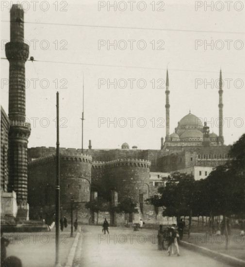 Port Said, View of a square in Port Said, stereograph, 1911, photographer, North Africa, paper, pasteboard, baryta paper, daylight gelatin silver print, height 90 mm x width 179 mm, square, circus, A monochrome scene showing a wide street leading toward a large stone fortress with rounded towers and crenellated walls, flanked on the left by a tall patterned column and on the right by a domed building with slender minarets rising above it; several vertical poles and wires cross the sky, trees line the right side of the street, and small groups of pedestrians and a few figures walking or standing are visible along the roadway.