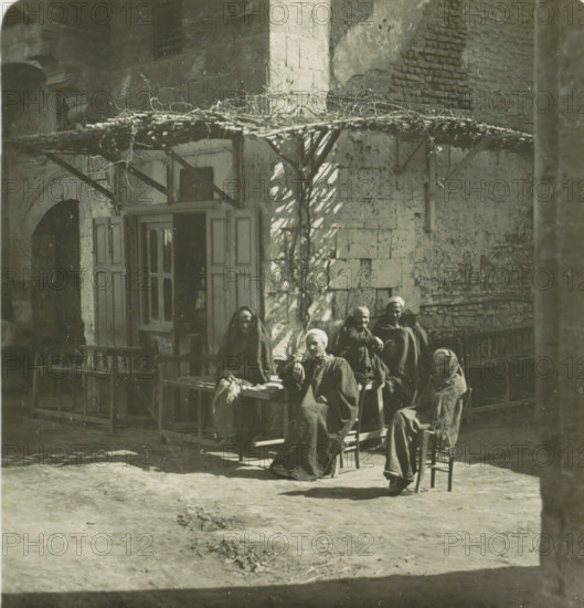 Luksor. Arabs Cafe, Guests in front of a café in Luxor, stereograph, number: '148', Luxor, publisher: Neue Photographische Gesellschaft, Steglitz, 1909 - 1935, photographer, North Africa, paper, baryta paper, gelatin silver print, developing gelatin silver print, height 88 mm x width 179 mm, pavement, terrace in front or inn or restaurant, An exterior scene shows a weathered stone building with a wooden door and shutters partly open beneath a simple pergola of branches and vines; in front of the building a group of people wearing dark, draped garments and head coverings sit and stand on benches and chairs, some leaning forward and some with arms crossed, their faces partially in shadow; the ground in front is bare earth with faint footprints and patches of darker material, and the sunlight casts irregular shadows from the pergola and nearby structures across the wall and ground.