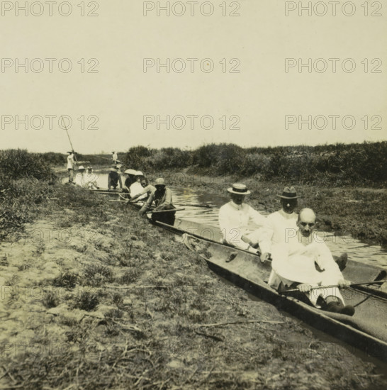 Some boats on a narrow canal, probably the Sungai Martapura river, with both Europeans and original inhabitants of Borneo, Netherlands. Indie, Borneo, with both whites and natives (former title), stereograph, Indonesia, 1900 - 1940, photographer, paper, pasteboard, baryta paper, gelatin silver print, developing gelatin silver print, height 90 mm x width 178 mm, river, A group of people sit in narrow wooden boats along a shallow waterway bordered by grassy banks and low shrubs, several individuals wearing light-colored clothing and hats, some straw boater hats and some wide-brimmed hats, with a few rowers holding oars and one person standing in the background using a long pole, the foreground showing rocky, grassy ground leading to the boats and water reflecting parts of the vessels and occupants.